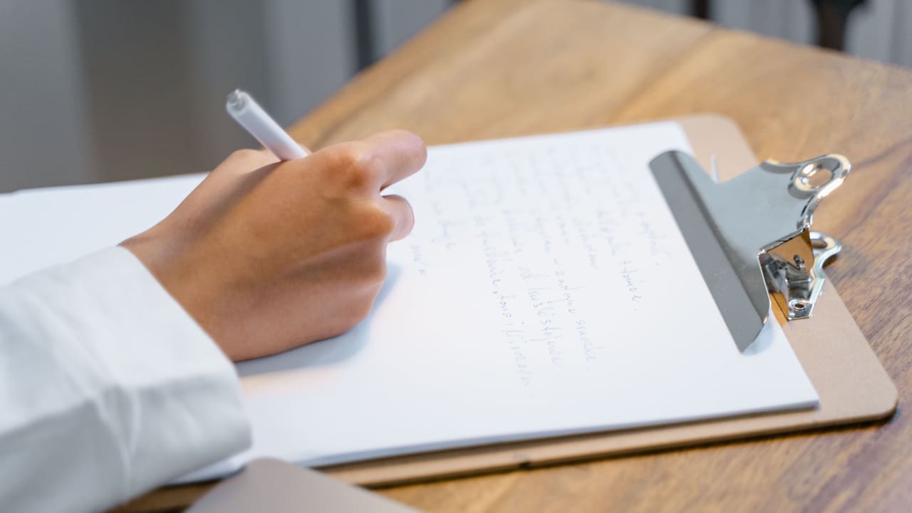 A hand writing on a clipboard on a wooden table.