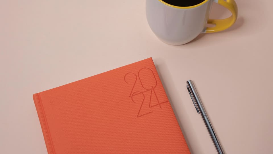 Stylish desk layout featuring a 2024 planner, cup of coffee, and pen on a light background.