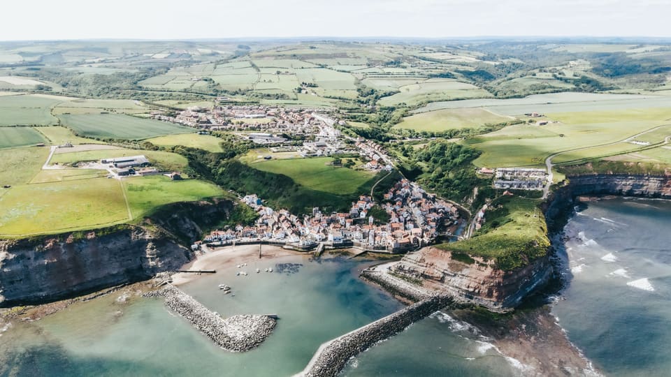Dramatic aerial shot of a coastal village surrounded by cliffs and green fields.