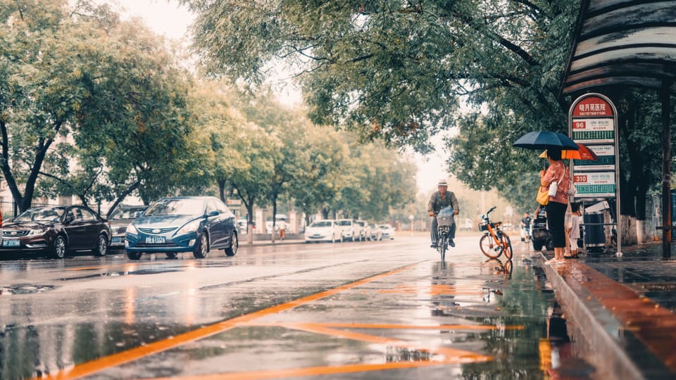 People wait under umbrellas at a Beijing bus stop while a cyclist rides by on a rainy day.