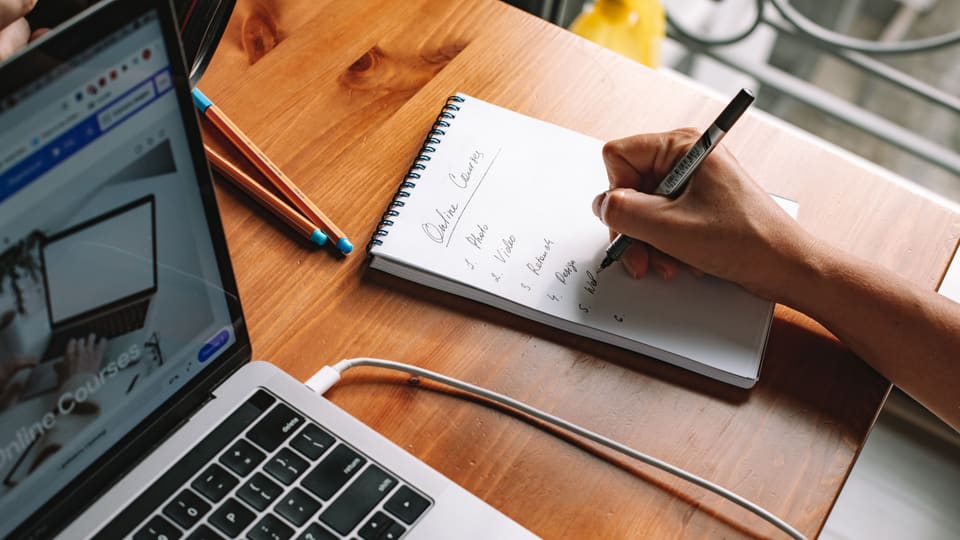 A person writing notes while researching online courses on a laptop in a home setting.